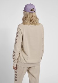 Felpa beige con maniche lunghe caratterizzata da motivi a zig-zag marroni sulle maniche; abbinata a pantaloni beige coordinati e un cappellino viola.