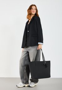Sac fourre-tout noir avec une texture lisse, deux longues anses et un petit nœud décoratif. Le modèle porte un blazer noir et un jean gris. Baskets argentées.