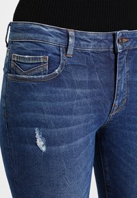 Jeans de mezclilla azul oscuro con un ajuste clásico, que presentan un detalle desgastado en el bolsillo delantero y costuras en contraste alrededor de la cintura.