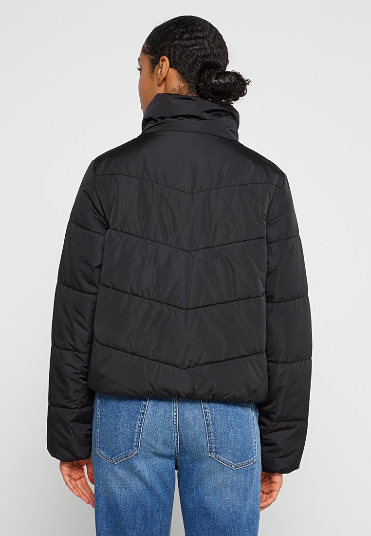 Giacca imbottita nera con colletto alto, caratterizzata da motivi di cucitura a zigzag e una vestibilità morbida. Indossata sopra pantaloni di jeans blu.