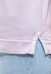 Χαμηλό μέρος πουλόβερ ανοιχτό μωβ με πλαϊνό άνοιγμα πάνω από ανοιχτό μπλε τζιν, εμφανίζοντας λεπτομέρειες ραφής και υφής υφάσματος.