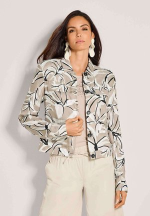 Vrouw draagt beige wijde broekspijpen en een beige bloes met bloemenprints en knopen, met grote witte oorbellen, staand tegen een egale achtergrond.