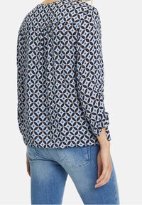 Blouse à manches longues avec un motif géométrique en bleu et marine. Tissu doux avec des détails froncés aux poignets. Associée à un jean clair.