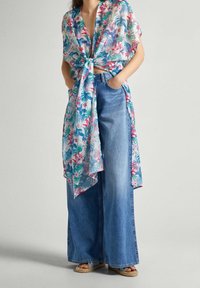 Kvinna som bär vida blåjeans och en knuten, kortärmad blommönstrad topp med händerna i fickorna och sandaler.