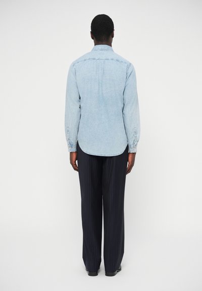 Hellblaues Jeanshemd mit langen Ärmeln, spitzem Kragen und abgerundetem Saum. Schwarze gestreifte Hose und dunkle Schuhe vervollständigen das Outfit.