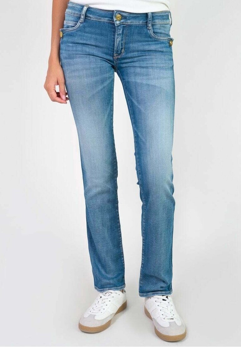 Femme portant un jean slim bleu clair et des baskets blanches, debout devant un fond uni.