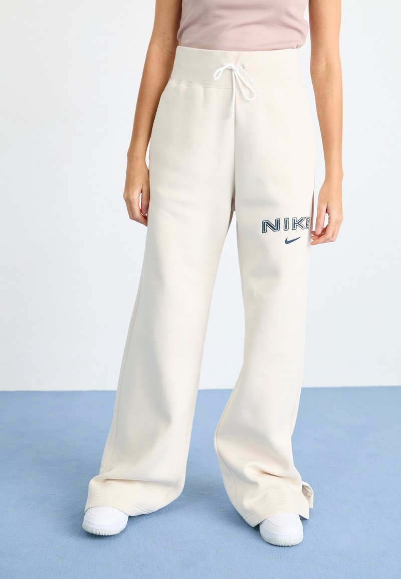 Svetle béžové tepláky s vysokým pasem a šňůrkou, se modrým logem a textem na levém boku. Široké nohavice s hladkým povrchem.