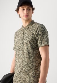 Grün-olivfarbenes Poloshirt mit Kurzarm, das mit einem schwarzen Blumenmuster, einem Kragen und drei Knöpfen versehen ist. Der Stoff wirkt glatt und leicht.