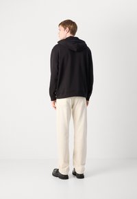 Sudadera negra con capucha y corte ajustado, combinada con pantalones holgados de color crema y zapatos negros, vista desde la parte trasera.
