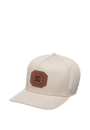 Beige honkbalpet met een bruine leren patch met de tekst "DC Shoe Company Quality Goods Est. 1994" op het voorpaneel.