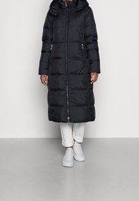 Sort pufferjakke med hætte, der har en frontlynlås, to sidelommer og quiltet tekstur. Bæret over hvide bukser og sneakers.