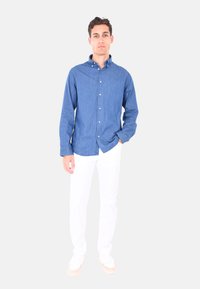 Camicia in chambray blu con colletto button-down e maniche lunghe, indossata con pantaloni bianchi e sneaker chiare, in piedi davanti a uno sfondo semplice.