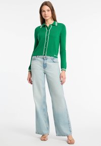 Zielony dzianinowy kardigan z białymi wykończeniami i guzikami, w połączeniu z jasnoniebieskimi dżinsami w stylu flare. Kardigan ma kołnierz i subtelne logo.