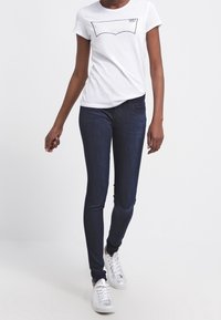 Camiseta blanca de manga corta con gráfico negro, jeans oscuros ajustados y zapatillas blancas. Diseño sencillo y silueta ajustada.