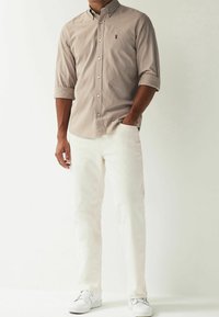 Gestreiftes Hemd zum Knöpfen in Hellbraun und Creme; lange Ärmel, hochgekrempelte Manschetten. Passende weiße Hose; einfarbige Textur. Weiße Sneakers.