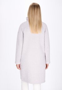 Donna con i lunghi capelli biondi, che indossa un cappotto grigio chiaro a maniche lunghe e pantaloni bianchi, di spalle contro uno sfondo bianco uniforme.