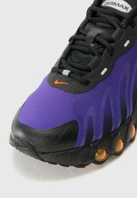 Sapatilha Nike Air Max com parte superior em malha roxa, detalhes em preto, sola de borracha texturizada e logo laranja. Materiais compostos e design em onda.