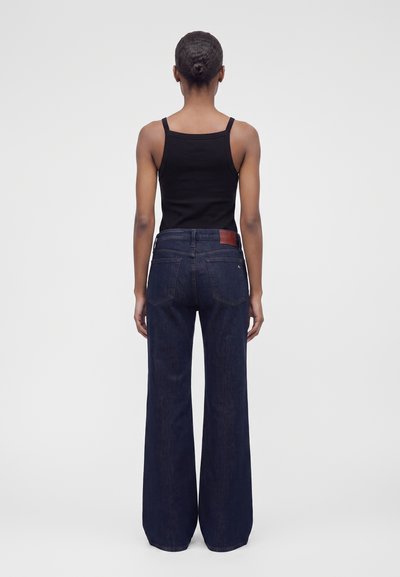 Femme aux cheveux en chignon, portant un haut noir sans manches et un jean large bleu foncé, debout de dos sur un fond gris uni.