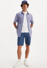 Short-sleeve, light blue linen shirt over a white T-shirt, paired with blue denim shorts and white sneakers. Blue hat completes the look.