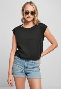 Černý top s krátkým rukávem a širokým výstřihem, doplněný světle modrými džíny s ohrnutými lemy. Modelka nosí tmavé sluneční brýle a visací náušnice.