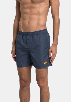 Torace e gambe maschili che indossano pantaloncini da bagno blu navy con fascia elastica in vita e una piccola toppa rossa e gialla sulla gamba sinistra.