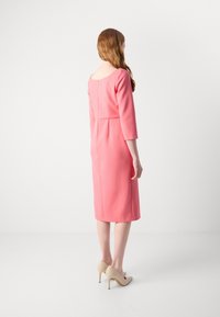 Robe rose ajustée avec des manches trois-quarts, un dos en forme de scoop et des détails de couture dans le dos, associée à des chaussures à talons hauts nude.