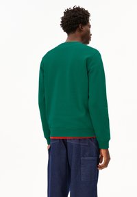 Felpa verde con scollatura rotonda e polsini a costine, caratterizzata da un colore uniforme e una texture liscia, indossata sopra una maglietta rossa.