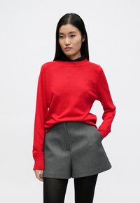 Suéter rojo de punto con cuello redondo acanalado y mangas largas, combinado con pantalones cortos grises entallados. La modelo lleva medias opacas negras.