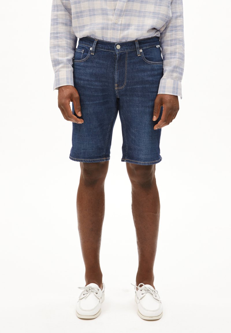 Donkere blauwe denim shorts, met een klassiek vijf-pocket ontwerp, rechte snit en opgerolde zoom. Gecombineerd met witte veterschoenen.