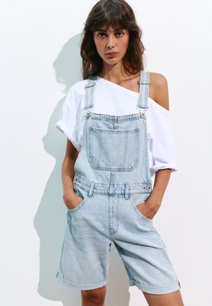 Jeune femme aux cheveux foncés portant une salopette courte en denim clair et un t-shirt blanc épaules dénudées, mains dans les poches, debout contre un mur blanc.