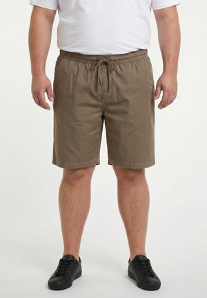 Uomo con pantaloncini marroni con coulisse, t-shirt bianca e scarpe da ginnastica nere, in piedi davanti a uno sfondo chiaro uniforme.