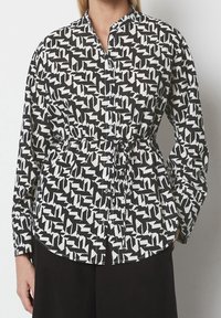 Camicia a motivi bianco e nero con colletto a band, chiusura con bottoni e vita cinchata per dare struttura. Maniche lunghe e orlo curvo.