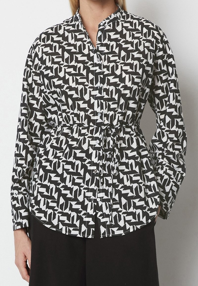 Camicia a motivi bianco e nero con colletto a band, chiusura con bottoni e vita cinchata per dare struttura. Maniche lunghe e orlo curvo.