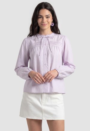Vrouw draagt een lichtpaarse blouse met lange mouwen en knopen en een witte rok, lacht met handen zachtjes voor zich geklapt.