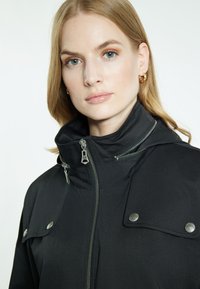 Primo piano di una donna con capelli biondi che indossa orecchini a cerchio dorati e un giubbotto nero con colletto alto e cerniere e bottoni in argento visibili.