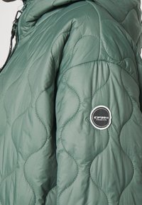 Manica di giacca trapuntata verde con patch circolare nero e bianco del logo Icepeak e dettagli della cerniera visibili vicino al colletto.