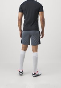 Chemise noire à manches courtes avec des accents blancs sur les côtés, shorts gris, chaussettes blanches montant jusqu'aux genoux et chaussures de football noir et rouge, vues de l'arrière.