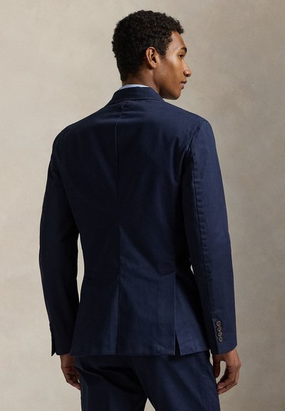 Muž viděn zezadu, oblečený v tmavě modrém saka s knoflíkovými detaily na manžetě, stojící na jednobarevném béžovém pozadí.