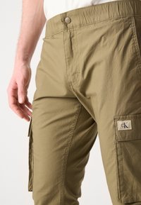 Olijfgroene cargobroek met een knoopsluiting, elastische tailleband, zijzakken en een klein merklabel op de bovenbeen. Lichte stofstructuur.