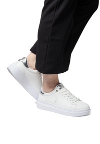 Zapatillas de cuero blancas con detalles plateados en el talón, que cuentan con una suela blanca gruesa, punta redondeada y un diseño con cordones. Logo detallado en el lateral.