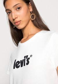 Camiseta blanca de algodón con un logo negro de "levi's". Presenta cuello redondo y mangas cortas. El modelo lleva pendientes de aro dorados.