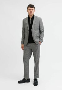 Completo grigio su misura con finitura strutturata, abbinato a un maglione nero con zip e scarpe nere. Design semplice e moderno.