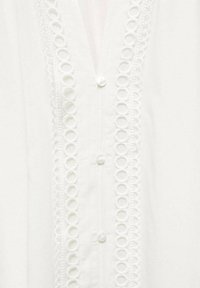 Witte blouse met een ronde kanten rand aan de voorkant, met standaard knoopsluitingen. Soepele stoftextuur en eenvoudig ontwerp.