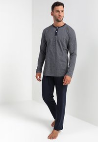 Férfi mezítláb áll, navy kék nadrágot és hosszú ujjú, navy és fehér csíkozott Henley pólót visel, háttérben egy fehér fal.