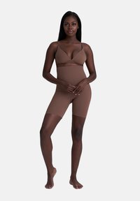 Vrouw in bruine hoge shapewear-shorts en bijpassende beha, staand op blote voeten tegen een effen lichte achtergrond.