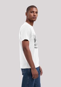 Witte katoenen T-shirt met een grafisch ontwerp op de voorkant. Model draagt een blauwe spijkerbroek, gezien vanaf de zijkant.