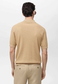Polo beige à côtes, avec des manches courtes et un col plat, présentant une texture subtile. Porté avec un pantalon beige ajusté.
