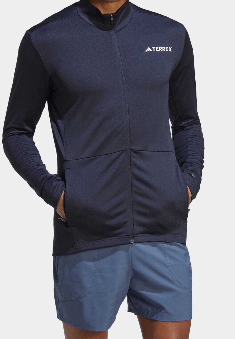 Granatowa kurtka z długim rękawem z zamkiem błyskawicznym, z dwoma przednimi kieszeniami, wykonana z rozciągliwego materiału, z haftowanym logo na lewej piersi.