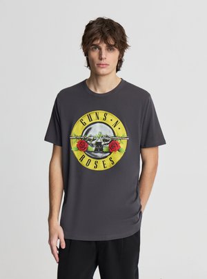 Młody mężczyzna z brązowymi włosami, ubrany w czarną koszulkę Guns N' Roses z czaszką i dwoma czerwonymi różami na żółtym okrągłym tle.