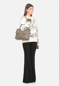 Donna in pantaloni neri a palazzo e maglione bianco con stampe grafiche nere, che tiene una grande borsa trapuntata taupe con dettagli dorati.